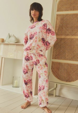 Cvetni pajamas set v mehkem roza materialu, ki ima velike vzorce vrtnic v odtenkih roza in zelene, z elastičnim pasom in ohlapnim krojem.