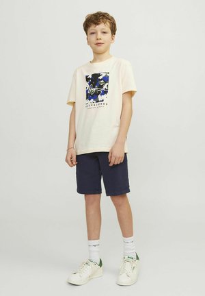 Ljust beige t-shirt med en blå och svart grafisk design, kombinerad med marinblå shorts och vita sneakers med gröna accenter.