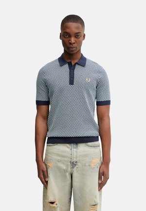 Hombre con camisa polo de manga corta a cuadros azul y blanca con cuello azul marino y vaqueros desgastados de lavado claro, de pie contra un fondo blanco.