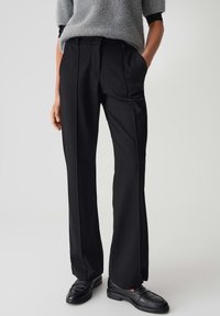Persoon in zwarte op maat gemaakte broek met voorplooien, zwarte loafers en een grijze fuzzy trui met korte mouwen, met de handen in de zakken.