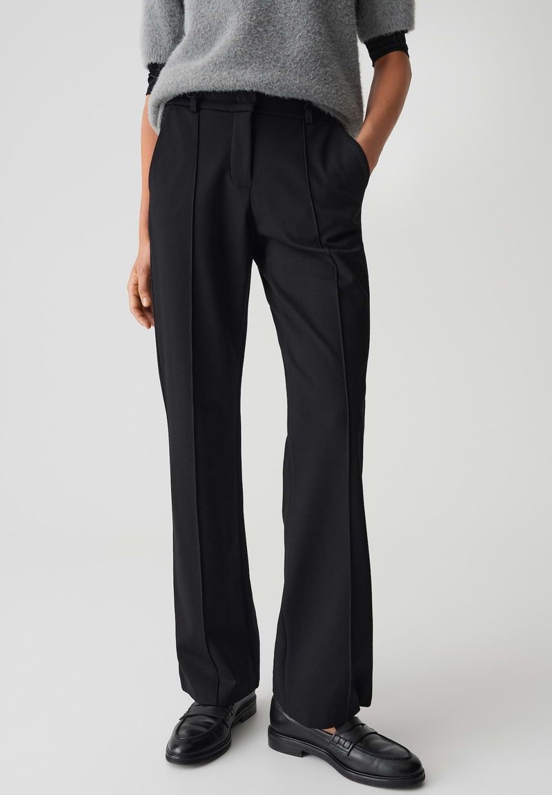 Persoon in zwarte op maat gemaakte broek met voorplooien, zwarte loafers en een grijze fuzzy trui met korte mouwen, met de handen in de zakken.