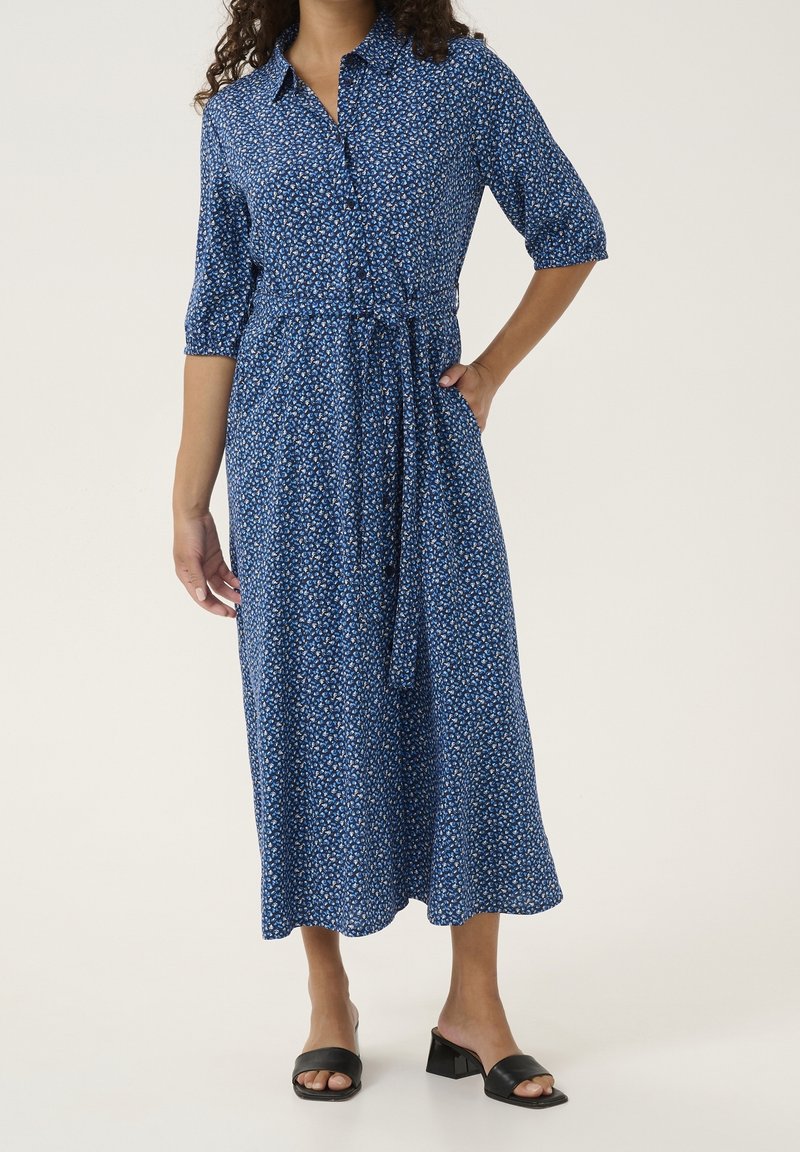 Femme portant une robe midi bleue à fleurs avec manches ¾, un col, des boutons et une ceinture à la taille, associée à des sandales à talons noires.