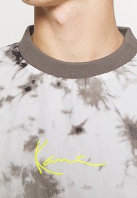 Camiseta de tirantes cortos, con estampado de tie-dye en tonos gris y blanco. Presenta un cuello acanalado gris y un logo bordado amarillo en la parte delantera.