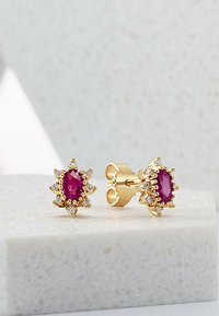 Goldene Ohrringe mit einem zentralen ovalen Rubin, umgeben von kleinen klaren Steinen, im floralen Design mit spitzen Akzenten.
