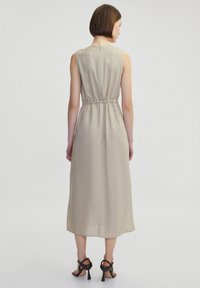 Abito smanicato di un beige chiaro con vita arricciata, lunghezza midi e texture liscia. Sono indossate sandali neri con tacco a stiletto e cinturini.