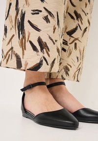 Ballerines en cuir noir avec un bout pointu et une bride à la cheville. Portées avec un pantalon beige présentant un motif de coups de pinceau brun et noir.