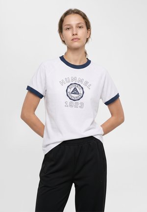 Ung kvinde med lysebrunt hår bærer en hvid T-shirt med navy kant og sorte bukser, står med hænderne bag ryggen.