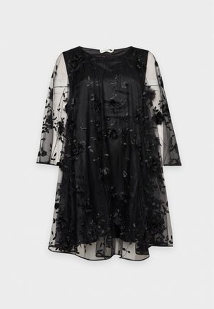 Un mannequin se tient devant un fond clair, portant une robe noire courte à manches transparentes à motifs floraux et des chaussures noires à talons hauts.