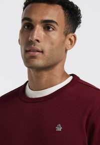 Jeune homme aux cheveux bouclés courts portant un sweat-shirt bordeaux avec un petit logo de pingouin sur la poitrine sur un t-shirt blanc à col rond.