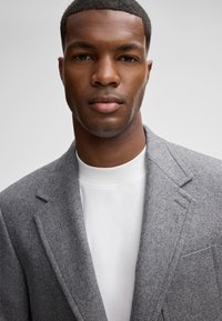 Grauer Wollblazer mit Reverskragen, strukturiertem Stoff und einer aufgesetzten Tasche über einem weißen Rundhals-Shirt. Nahaufnahme mit neutralem Hintergrund.