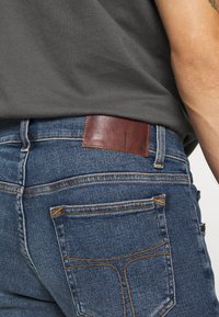 Jeans i denim med mörkblå färg, med orange sömmar, en läderpatch på baksidan med logotyp och en slim-fit design.