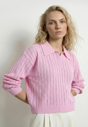 Ung kvinde med bølget blondt hår iført en pink strikket langærmet sweater og lysfarvet plisseret nederdel mod en ensfarvet baggrund.