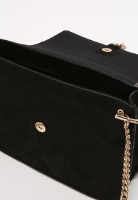 Borsa a mano in suede nero con tracolla a catena dorata, forma rettangolare elegante e chiusura semplice a pulsante. Interno non foderato.