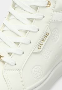 Sneakers sintetiche bianche con logo in rilievo e motivi circolari, dotati di lacci bianchi e dettagli in hardware dorato sul lato.