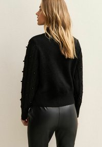 Donna con lunghi capelli biondi, che indossa un maglione nero con maniche strutturate e pantaloni di pelle nera, in posa di profilo contro un muro semplice.