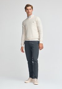 Kremowy sweter z dekoltem w kształcie litery V, wykonany z miękkiego materiału, w połączeniu z ciemnoszarymi spodniami i jasnobeżowymi sneakersami. Dyskretne logo na klatce piersiowej.