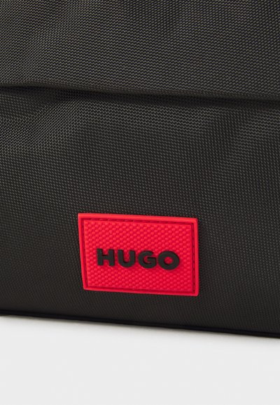 Sac en tissu noir avec une surface texturée et un logo carré rouge bien en vue avec "HUGO" en lettres noires.