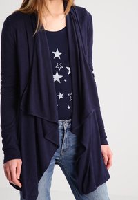 Mörkblå cardigans med mjukt material, med en lager-på-lager design framtill. Matchas med en mörk topp med stjärnmönster och blå jeans.