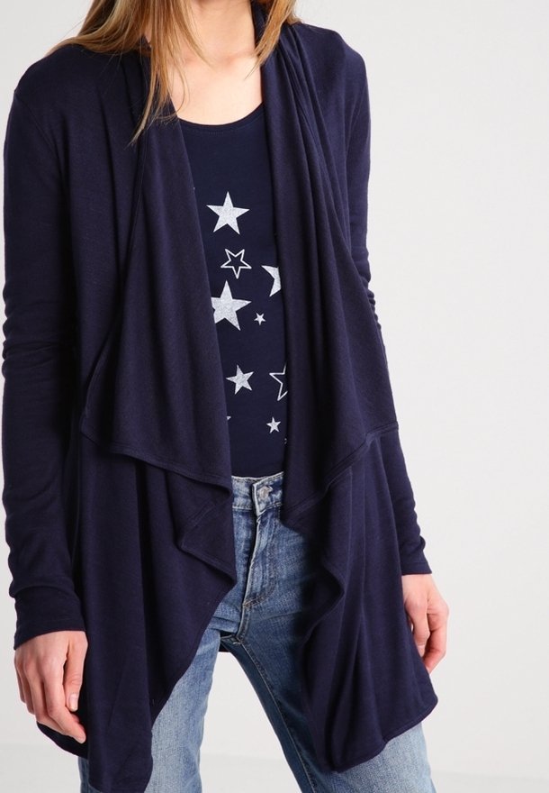 Mörkblå cardigans med mjukt material, med en lager-på-lager design framtill. Matchas med en mörk topp med stjärnmönster och blå jeans.