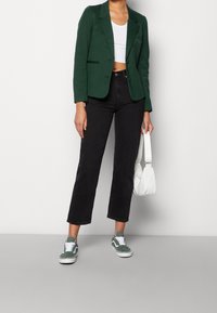 Zelený blazer přes bílý crop top, doplněný černými džínami s vysokým pasem a zelenými teniskami. Bílá taška, hladký materiál, přiléhavý střih.