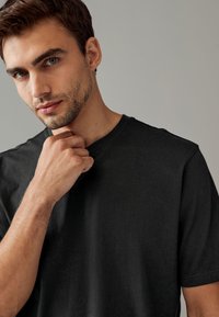 T-shirt nera a maniche corte con vestibilità comoda, caratterizzata da scollo a V e tessuto morbido e liscio. Modello posa con la mano sotto il mento.