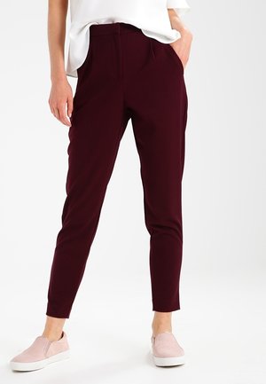 Persona con pantalones ajustados de color burdeos oscuro y zapatos tipo slip-on rosa claro, de pie con una mano en el bolsillo.