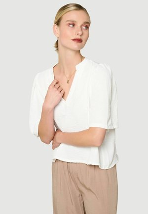 Frau mit blondem Haar in einer weißen Bluse und beigen Hosen, die seitlich blickt, mit einer Hand am Schlüsselbein und der anderen über der Taille verschränkt.