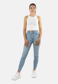 Woman standing wearing a white sleeveless crop top, light blue high-waisted jeans, white sneakers, and gold dangling earrings.