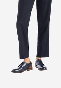 Chaussures en cuir noires avec un accent en cuir marron sur le talon ; elles possèdent des lacets, un bout rond et une texture lisse. Associées à un pantalon sombre ajusté.