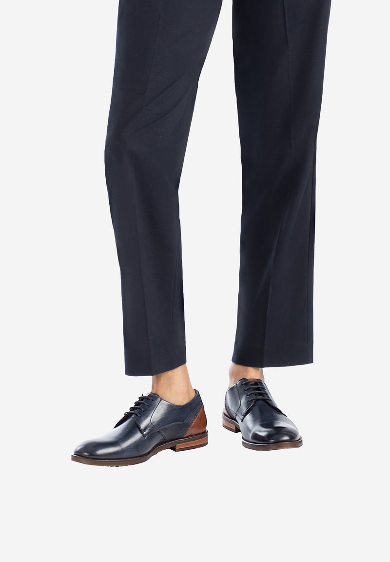 Chaussures en cuir noires avec un accent en cuir marron sur le talon ; elles possèdent des lacets, un bout rond et une texture lisse. Associées à un pantalon sombre ajusté.