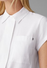Camicia bianca a maniche corte con colletto button-down, dotata di una tasca sul petto a sinistra e di un piccolo logo argento. Tessuto dalla superficie liscia.