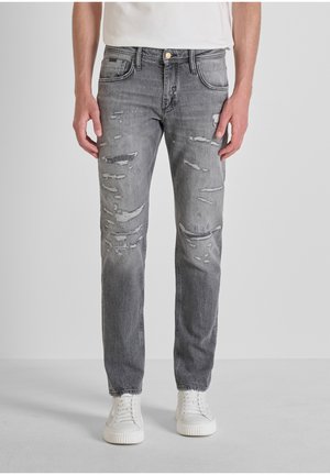 Man in grijze versleten spijkerbroek en witte sneakers, staand tegen een effen lichte achtergrond.