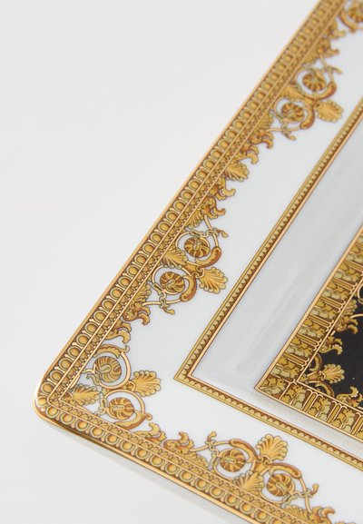 Dekoratívny porcelánový tanier so zlatým ozdobným okrajom, zdobený zložitými listovými a kvetinovými motívmi, prevažne biely s čiernymi akcentmi.