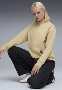 Beige zip-up sweatshirt med kengurulomme og broderet logo, parret med mørke vide bukser og hvide sneakers foran en grå baggrund.