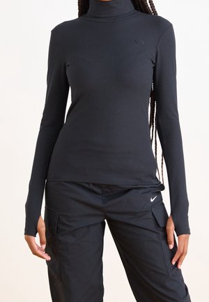 Femme portant un col roulé côtelé noir et un pantalon cargo noir Nike avec des logos de marque visibles sur les deux vêtements.