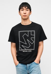 Μαύρο βαμβακερό T-shirt με λευκό γεωμετρικό λογότυπο και κείμενο «MICHAEL KORS» στο κάτω μέρος. Κοντά μανίκια, στρογγυλή λαιμόκοψη.