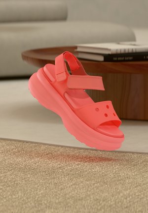 Jasno koralowa sandałka na platformie z regulowanymi paskami unosząca się nad beżowym dywanem, owalnym drewnianym stolikiem kawowym i stosami książek w tle.