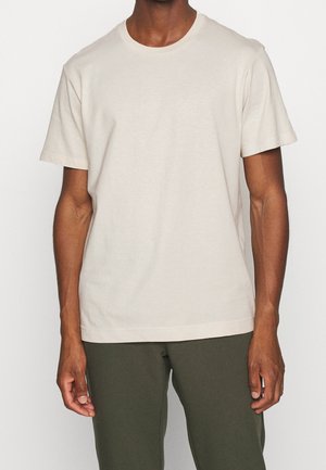 Mężczyzna ubrany w gładki, jasnobeżowy t-shirt z krótkim rękawem i ciemnozielone spodnie, stojący na neutralnym tle.