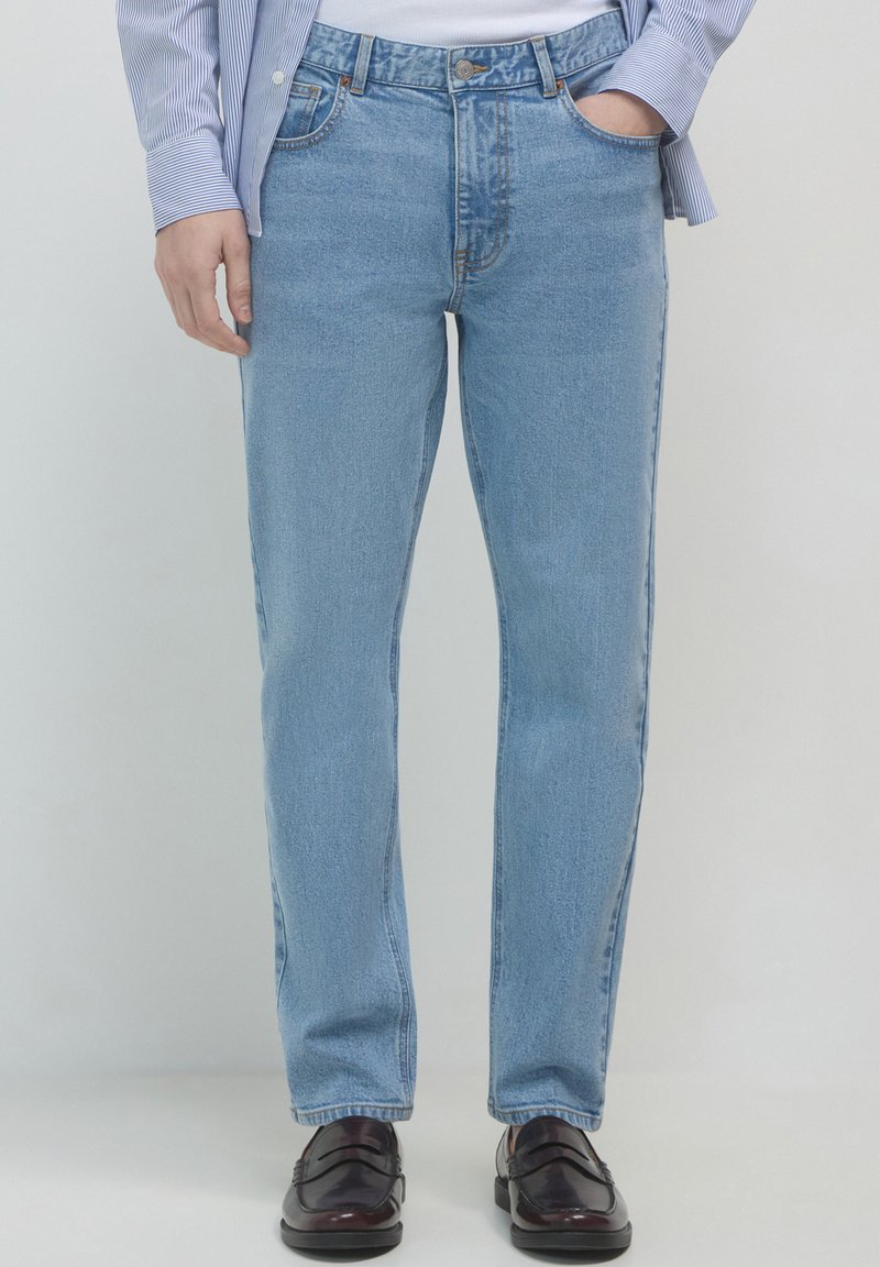 Persona che indossa jeans a gamba dritta di colore azzurro chiaro con una mano in tasca e mocassini in pelle marrone scuro su uno sfondo semplice.