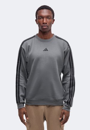 Άνδρας φοράει γκρι φούτερ Adidas με μαύρες ρίγες στα μανίκια και μπεζ παντελόνι, στέκεται μπροστά σε ανοιχτό φόντο.