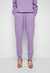 Fialové joggingové kalhoty vyrobené z bavlněné směsi s elastickými manžetami, nastavitelnou šňůrkou na stažení a bočními kapsami. V kombinaci s černými páskovými podpatky.