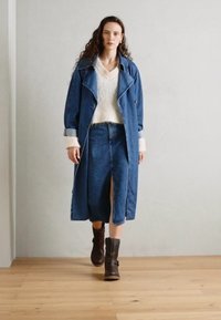 Denim-Mantel mit großen Revers, kombiniert mit einem flauschigen weißen Pullover und einem knielangen Jeansrock. Braune Stiefeletten runden den Look ab.