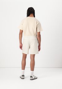 Beige T-Shirt und hellfarbene Shorts mit elastischem Bund, getragen mit weißen Socken und schwarz-weißen Sandalen. Rückansicht.