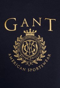 Granatowy dzianinowy materiał zdobiony złotym haftem z logo "GANT" oraz herbem z liśćmi laurowymi, zawierającymi napis "American Sportswear" i rok założenia.