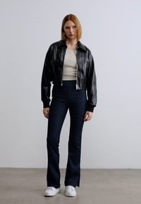 Μαύρο cropped δερμάτινο μπουφάν πάνω από μπεζ τοπ και σκουρόχρωμα τζιν καμπάνα, σε συνδυασμό με λευκά αθλητικά παπούτσια, σε ουδέτερο φόντο.