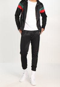 Svart idrottsoverall med röda och gröna ränder på ärmarna och sidorna, tillverkad av glänsande tyg med en kroppsnära passform. Vita sneakers på fötterna.