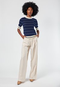 Navy striped knit top with cream accents, paired with beige high-waisted pleated trousers and tan pointed shoes. Smooth textures throughout.