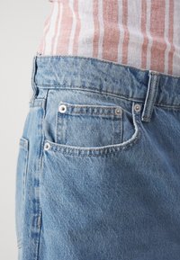 Jean en denim bleu avec détails aux poches avant, porté avec une chemise rayée rose et blanche rentrée à l’intérieur.