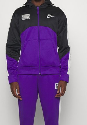 Mężczyzna w fioletowo-czarnym dresie Nike z białymi napisami "Force" i logo Nike, stojący na jednolitym tle.