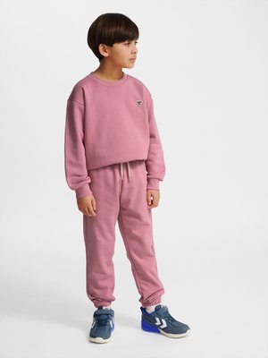 Dreng står iført lyserød sweatshirt og joggingbukser med blå og hvide sneakers, ser til siden på ensfarvet lys baggrund.